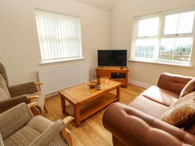 A living room with a brown sofa two armchairs a wooden coffee table with a bottle and two glasses and a television at Coquet in Longframlington near Alnwick