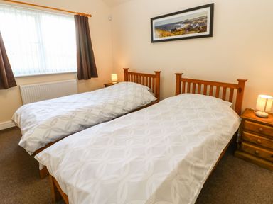 A bedroom with two single wooden beds white patterned bedding bedside tables with lamps a window with brown curtains and a landscape picture on the wall at Coquet in Longframlington near Alnwick