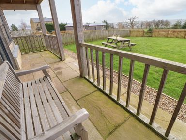 A wooden bench on a covered stone patio overlooking a fenced grass yard with a picnic table at Coquet in Longframlington near Alnwick
