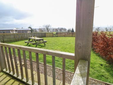 A fenced backyard with a wooden picnic table and benches viewed from a wooden porch at Coquet in Longframlington near Alnwick