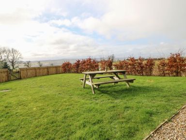 A wooden picnic table with two glasses and a bottle on a grass lawn with a wooden fence and trees in the background at Coquet in Longframlington near Alnwick