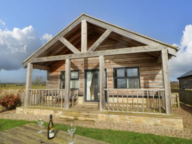 A wooden cabin with a porch bench and table with a wine bottle and glasses on the grass at Coquet in Longframlington near Alnwick