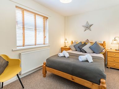 A bedroom with a wooden bed with pillows and towels a yellow chair a window with blinds and wooden bedside tables with lamps at Dringarth in Tenby