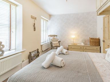 A bedroom with a bed covered with a quilt and rolled towels on top a wooden chair with cushions a dresser with a lamp and fan and two windows with blinds at Dringarth in Tenby