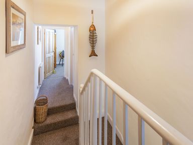 A hallway with stairs carpeted floor a basket on the floor framed pictures on the wall and a wooden fish hanging near a doorway at Dringarth in Tenby