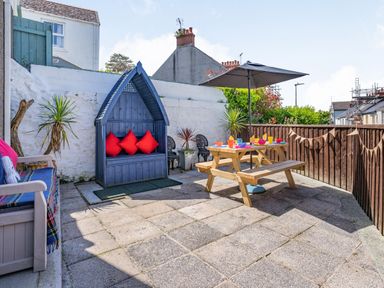 A paved outdoor patio with wooden seating including a bench with red cushions a picnic table with an umbrella and colorful tableware at Dringarth in Tenby