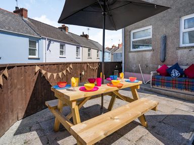 An outdoor patio with a wooden picnic table set with colorful bowls and plates and a large umbrella with a cushioned bench against a wall at Dringarth in Tenby