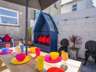 An outdoor dining area with a wooden table set with colorful bowls and cups a blue wooden canopy seat with red cushions and black metal chairs against a white wall at Dringarth in Tenby