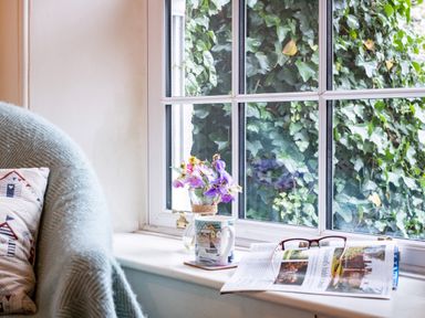 A window seat with a mug a vase of flowers an open newspaper and glasses next to a chair with a blanket and cushion at Cobble Cottage in Boscastle near Tintagel