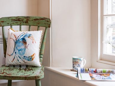 A distressed green wooden chair with a crab cushion beside a windowsill with a mug and an open magazine at Cobble Cottage in Boscastle near Tintagel