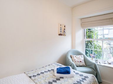 A bedroom with a bed covered in a patterned quilt and towels a green armchair with a cushion by a window with blinds and a small vase of flowers on the windowsill at Cobble Cottage in Boscastle near Tintagel