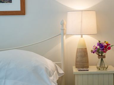 A bedside table with a lamp and a vase of flowers next to a bed with white bedding at Cobble Cottage Boscastle near Tintagel
