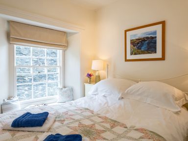 A bedroom with a bed covered by a quilt and towels near a window with cushions and a bedside table with a lamp and flowers at Cobble Cottage Boscastle near Tintagel