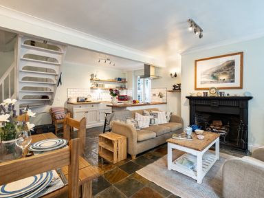 An open-plan living area with a kitchen, dining table, sofa, fireplace, and staircase at Cobble Cottage in Boscastle near Tintagel