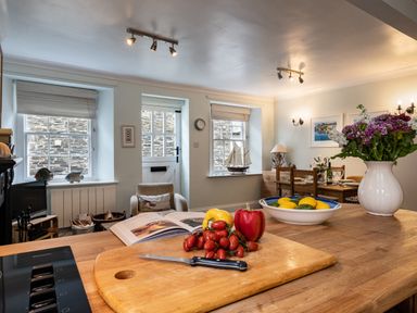 A kitchen island with a cutting board holding tomatoes and peppers a bowl of lemons and limes a flower vase a dining table set with chairs and a model ship on a windowsill at Cobble Cottage in Boscastle near Tintagel