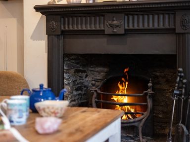 A fireplace with a fire burning and a table with a blue teapot and cups in front at Cobble Cottage in Boscastle near Tintagel