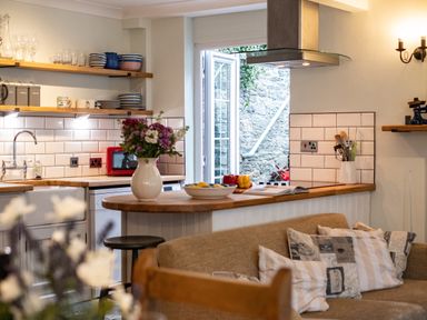 A kitchen with open shelves holding dishes and glassware a wooden countertop with a vase of flowers and stools a couch with pillows and a door leading outside at Cobble Cottage in Boscastle near Tintagel