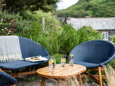 An outdoor seating area with blue wicker chairs a wooden table with glasses of water and a blanket on a chair at Cobble Cottage in Boscastle near Tintagel