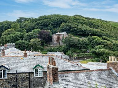 Roofs of stone houses with chimneys and a large brick house on a hill with trees in the background at Cobble Cottage in Boscastle near Tintagel