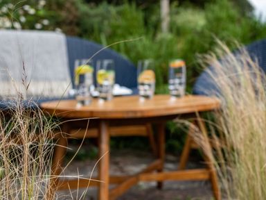 A wooden outdoor table with four glasses of water with lemon slices on a patio with rattan chairs and grass surrounding at Cobble Cottage Boscastle near Tintagel