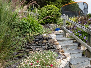 A garden with stone steps wooden railing flowers and shrubs with a trampoline in the background at Cobble Cottage in Boscastle near Tintagel
