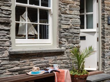A stone exterior wall with a window displaying a model sailboat a white door a bench with a hat a magazine a mug and a tablecloth and a potted plant at Cobble Cottage Boscastle near Tintagel