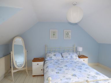 A bedroom with a bed and floral bedding two nightstands a standing mirror and two framed pictures on a blue wall at Woodbine Cottage in Old Leighlin near Kilkenny County Kilkenny