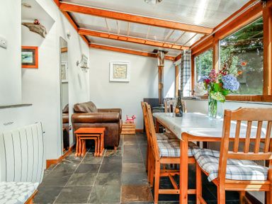 A dining area with wooden framed chairs and a table with flowers and wine glasses a leather armchair and a stove in the corner at Hwylfa'r Groes in Talybont near Barmouth