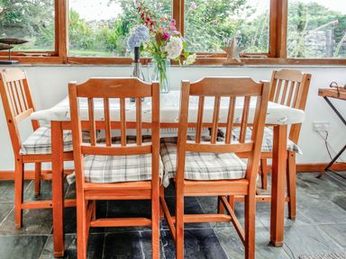 A dining table with a floral arrangement and four wooden chairs with plaid cushions in a room with large windows at Hwylfa'r Groes in Talybont near Barmouth