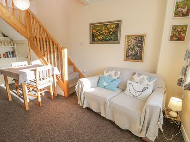 A living room with a wooden staircase a sofa with cushions a table with chairs and paintings on the walls at Bryn Melyn Artist's Cottage Llanelltyd near Dolgellau