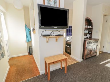 A room with a wall mounted television a wooden bench a hallway with a jacket hanging an antique cabinet with shelves displaying dishes and a view into a kitchen with an oven at Bryn Melyn Artists Cottage in Llanelltyd near Dolgellau