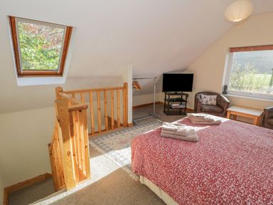 A bedroom with a red bedspread, wooden staircase railing, two armchairs, a small table, clothes rack, and a television at Bryn Melyn Artist's Cottage Llanelltyd near Dolgellau