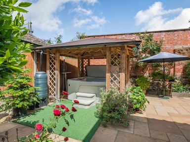 An outdoor patio with a hot tub under a wooden gazebo surrounded by plants and a table with an umbrella at The Lodge in Coltishall