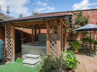 An outdoor area with a hot tub under a wooden gazebo surrounded by plants and a patio table with an umbrella at The Lodge in Coltishall