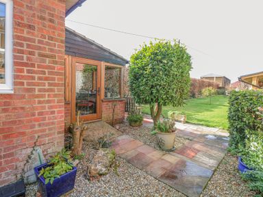 A garden with a paved path a pruned tree multiple potted plants and shrubs beside a house with wooden framed glass door at The Lodge in Coltishall