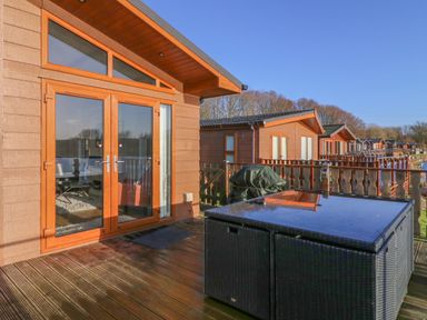 A wooden deck with a rattan table and glass door entrance at Shore Beats Work - Capenwray 10 in South Lakeland Leisure Village