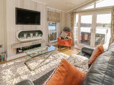 A living room with a leather sofa with cushions a glass coffee table an orange armchair a wall-mounted TV and a fireplace with candles at Shore Beats Work - Capenwray 10 in South Lakeland Leisure Village