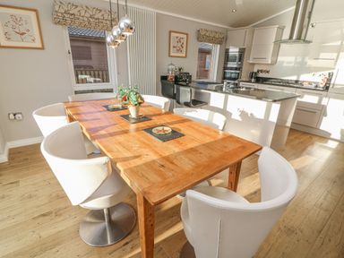 A dining area with a wooden table surrounded by white chairs next to a kitchen island with black stools at Shore Beats Work - Capenwray 10 in South Lakeland Leisure Village