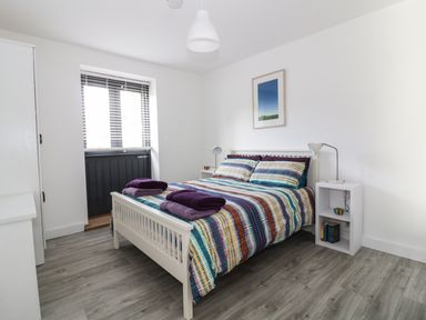 A bedroom with a bed covered in a multicolored striped duvet and purple towels on top two small white bedside tables one with a lamp and the other with books and a lamp and a window with blinds at Whitehands Farm Barn in Old Buckenham