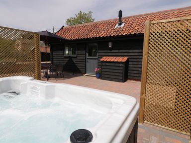 An outdoor patio area with a hot tub a table and chairs with an umbrella black wooden building with a tiled roof at Whitehands Farm Barn in Old Buckenham
