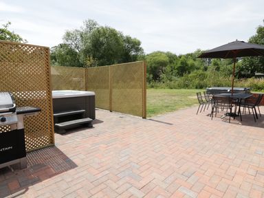 A patio with a hot tub behind wooden lattice screens a barbecue grill and an outdoor table with chairs and a large umbrella at Whitehands Farm Barn in Old Buckenham