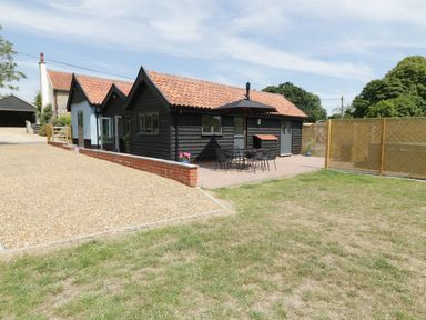 A small house with a red tiled roof and black wooden exterior with a patio table and chairs under an umbrella on a paved area at Whitehands Farm Barn in Old Buckenham