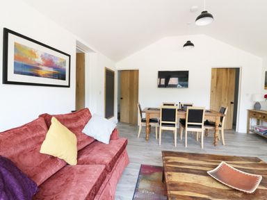 A living room with a red sofa and cushions a wooden dining table with chairs a wall-mounted tv and framed artwork at Whitehands Farm Barn in Old Buckenham