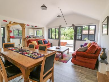 A dining area with wooden table and chairs next to a living room with red sofas and a coffee table at Whitehands Farm Barn in Old Buckenham