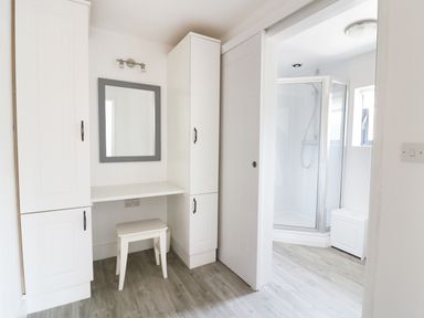 A dressing area with white cabinets a stool and a mirror next to a bathroom with a shower enclosure at Whitehands Farm Barn in Old Buckenham