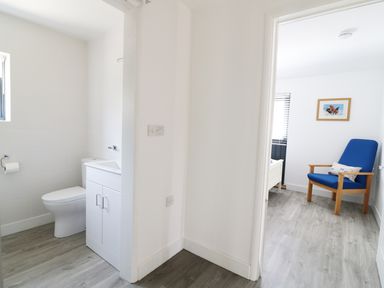 A hallway showing a bathroom with a toilet and sink on the left and a room with a blue armchair and framed picture on the right at Whitehands Farm Barn in Old Buckenham