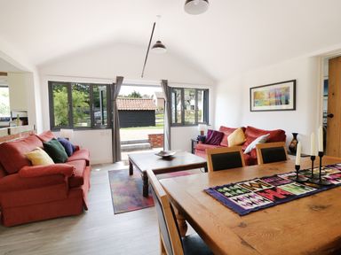 A living room with red sofas and colorful cushions near large windows and wooden dining table with candles at Whitehands Farm Barn in Old Buckenham