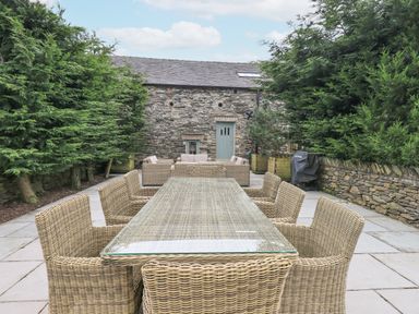 An outdoor patio with a glass top dining table and wicker chairs surrounded by tall trees at Yew Cottage in Backbarrow