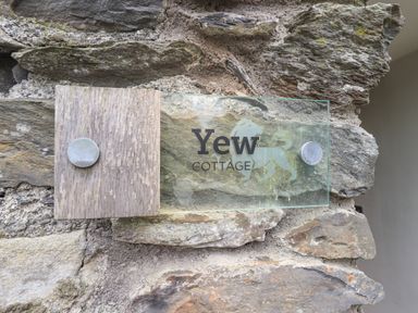 A glass and wood sign attached to a stone wall reading Yew Cottage at Backbarrow