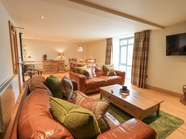 A living room with brown leather sofas green and plaid cushions wooden coffee table dining table and chairs with plaid curtains at Yew Cottage Backbarrow
